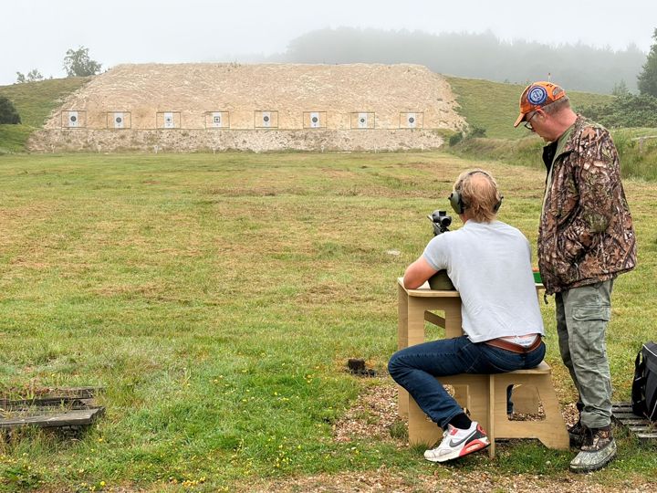 A member shooting from a bench with another member standing beside them, and the target butts behind