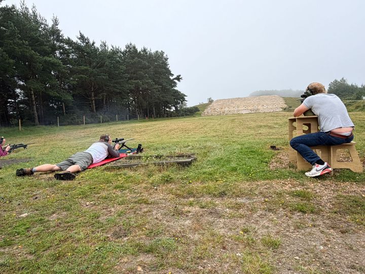 One member lying prone and another shooting from a bench, with pine trees behind