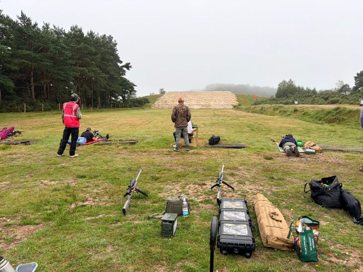 Rifle cases and equipment on the grass, with members and the target butts in the distance